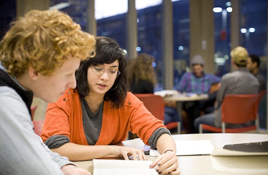 A female student sits with a male student, helping with him with his homework. The woman's hand is on the man's notebook, following a line of text as she reads.