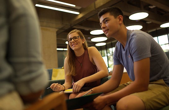 Two students are listening to a third student during a conversation, with one of the listening students taking notes on a pad of paper.