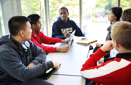 A group of students sits in a meeting room, having a discussion about a project.  One student is typing on a laptop while the student at the head of the table is speaking.