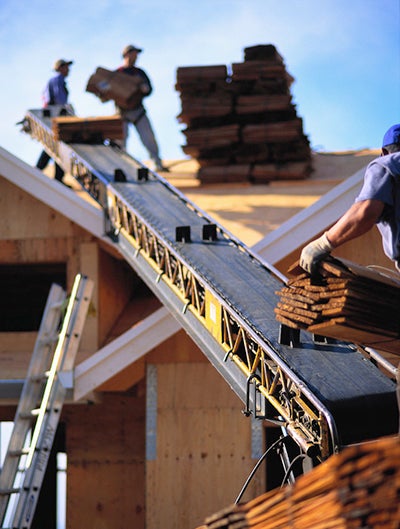 A construction worker sends a bundle of shingles up a conveyor belt toward a roof, where two workers are unloading the bundles of shingles and stacking them on the roof.