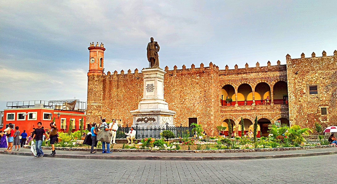 A plaza in Cuernavaca, Mexico, with a large statue standing in the middle of a brick-lined roundabout and people milling around the plaza.