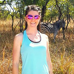 Alyson Klausing poses for a photograph with a zebra standing in a field in the background.
