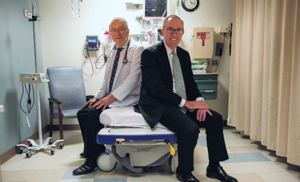 A hospital administrator poses for a photo sitting on a hospital bed with a doctor also sitting on the bed.