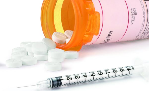 A graphic image showing a bottle of pills laying on its side, with pills spilled on the surface, and a needle laying along side.  The image represents the ongoing opioids epidemic.