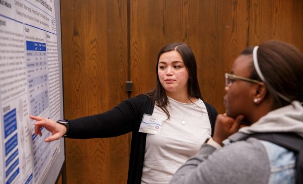 An SPH student presents her research while pointing to her research poster, and another student is listening to the presentation.