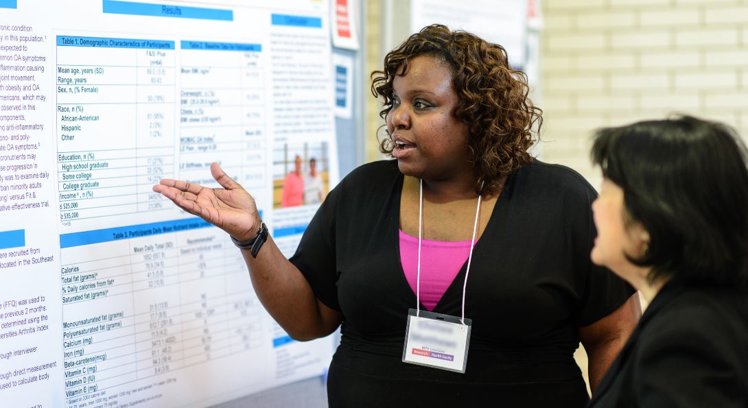 A student gestures at her poster presentation, explaining her research to an SPH staff member.