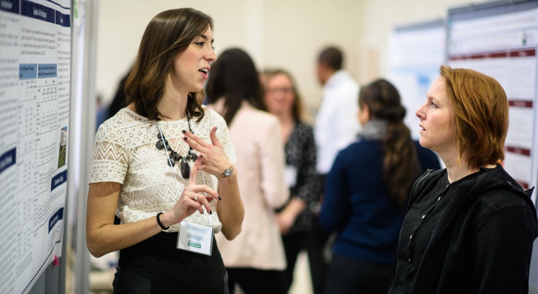 A student talks to another student while explaining her research poster presentation.