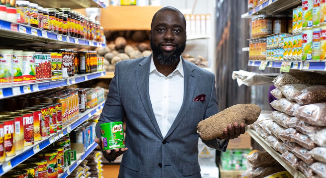 MHA alumnus Sobitan Boyede stands in a grocery aisle, holding a yuca and a canned good product.