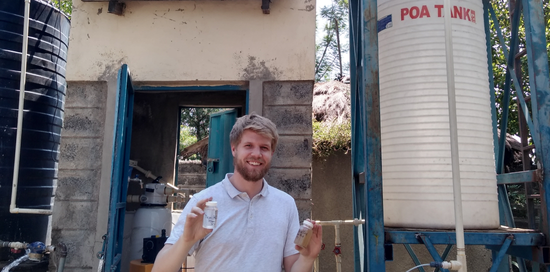 MPH student Colin Hendrickson poses for a picture in front of a solar powered water disinfection station in Kenya, holding up two vials of water.