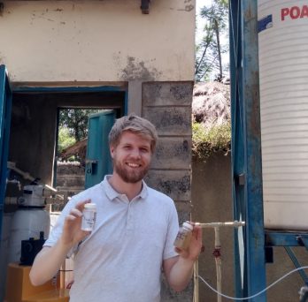 Colin Hendrickson poses for a photo holding two vials of cleaned water, standing in front of a water pumping station in Kisumu, Kenya.
                  