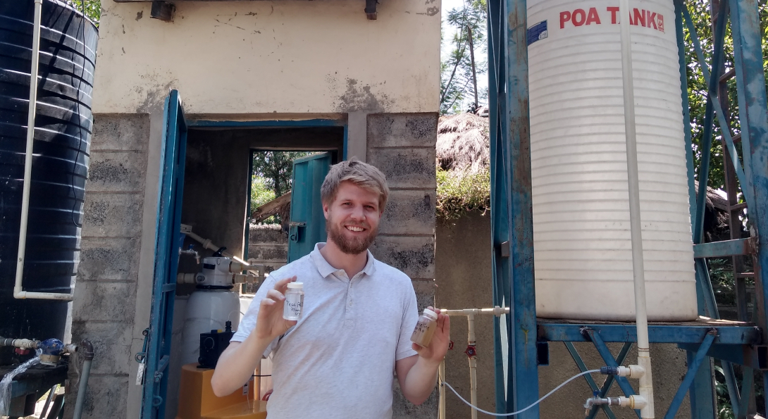 Colin Hendrickson poses for a photo holding two vials of cleaned water, standing in front of a water pumping station in Kisumu, Kenya.