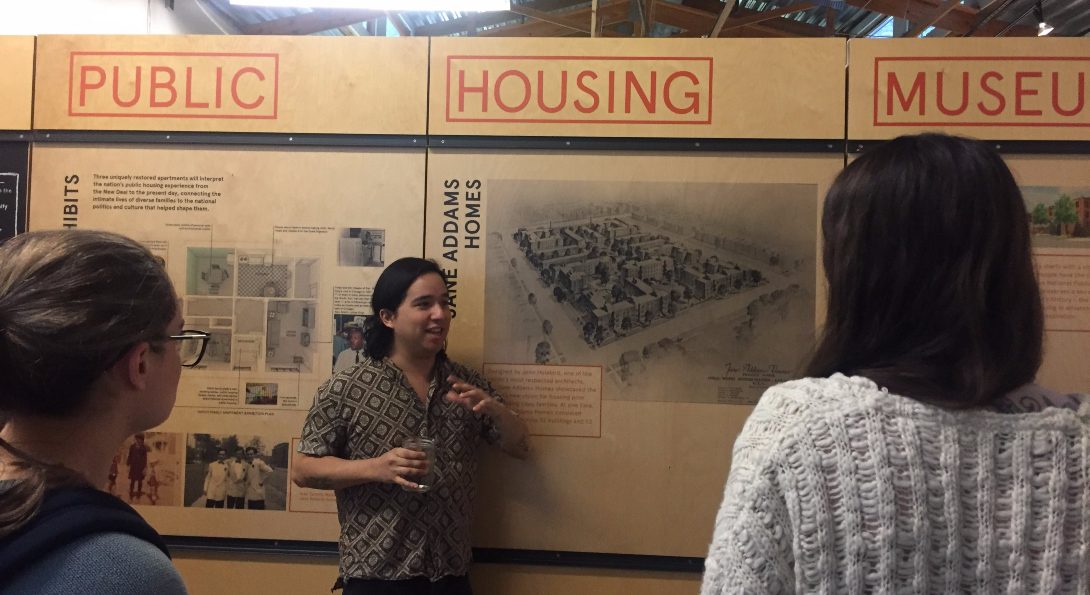 Students who took the Epidemics of Injustice course visit the National Public Housing Museum, listening to a presentation in front of a large poster on the history of public housing.