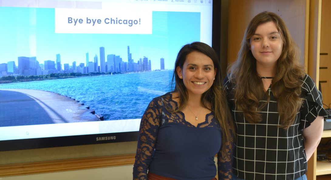 Students Vania Lara and Sara Izquierdo pose for a picture standing in front of a television screen with a presentation about their global experiences displayed.