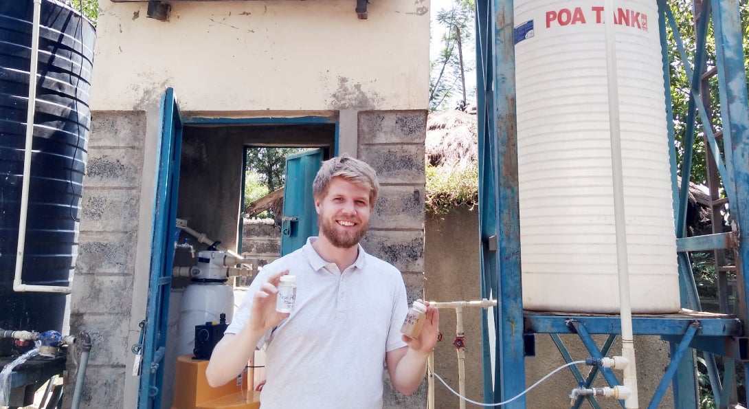 Colin Hendrickson, MPH student in environmental and occupational health sciences, stands by the water tanks he used for his project. In his hands are samples of the dirty river water and the clean water he disinfected.