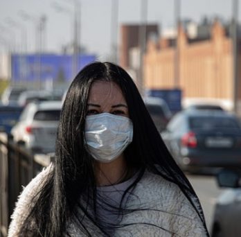 A woman wears a surgical mask while posing for a picture next to a busy street. 