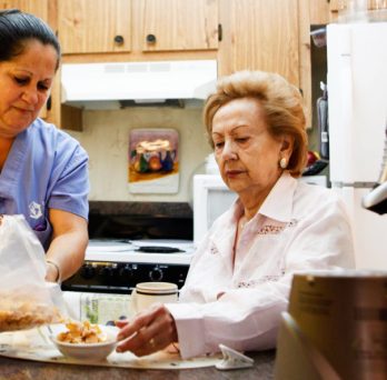 A home care aide prepares breakfast for an elderly woman in the woman's kitchen.
                  