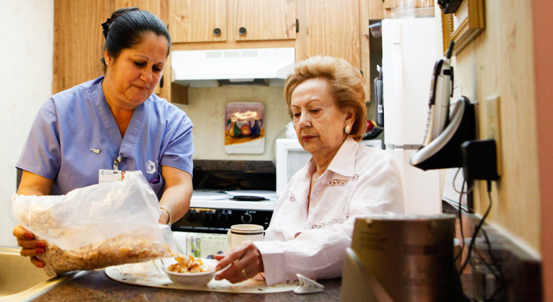 A home care aide prepares breakfast for an elderly woman in the woman's kitchen.