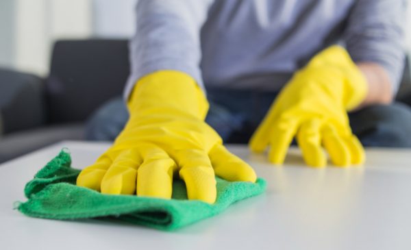 A person disinfects a table in their home with a green rag, while wearing yellow protective rubber gloves.