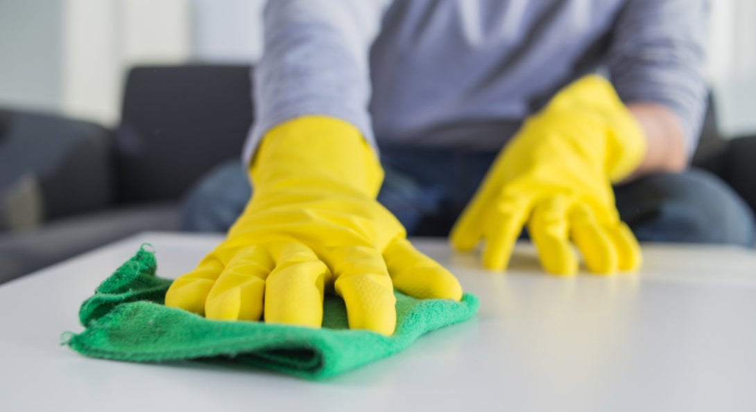 A person disinfects a table in their home with a green rag, while wearing yellow protective rubber gloves.