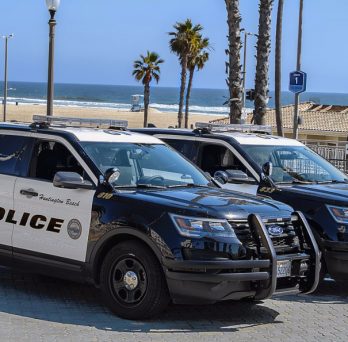 Two Huntington Beach police cars are parked near a beach, where police are monitoring beach closures during the COVID-19 pandemic.
                  
