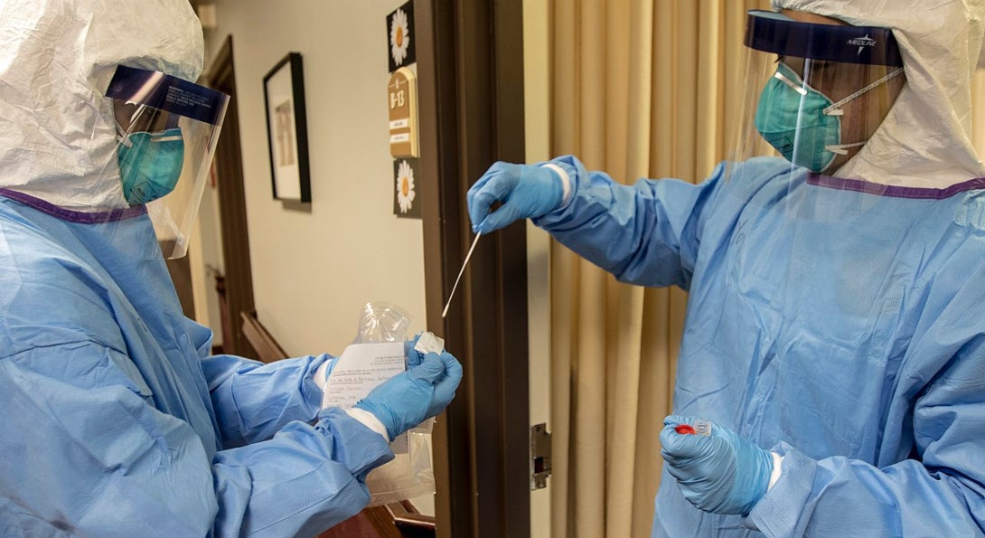 Two medical professionals, dressed in personal protective gowns, masks and face shields, prepare a COVID-19 test sample for processing.