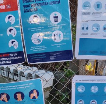 Various warning and safety signs are posted on a fence outside a construction site during the COVID-19 pandemic. 
