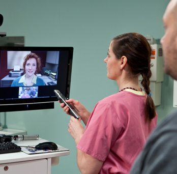 A telehealth consultation at a VA clinic, with a nurse adjusting a television set with a video feed from a doctor, as a patient waits in the background.
                  