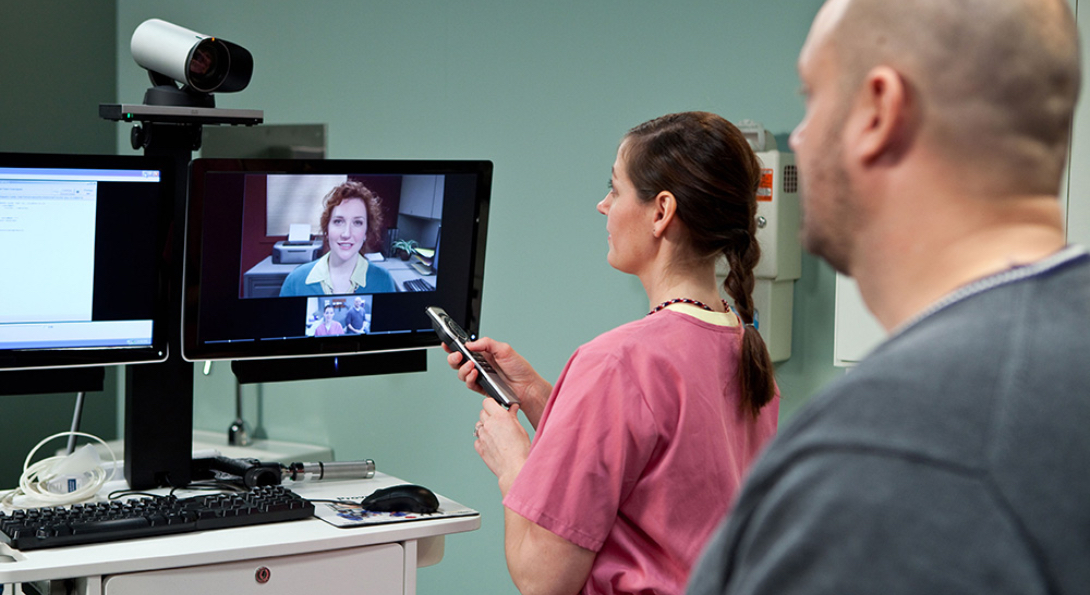 A telehealth consultation at a VA clinic, with a nurse adjusting a television set with a video feed from a doctor, as a patient waits in the background.
