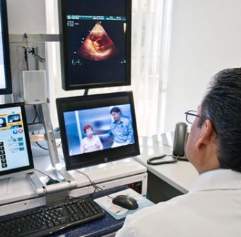 A doctor conducts a telemedicine consultation, viewing a screen with a video link to a patient and another computer screen with diagnostic information. 