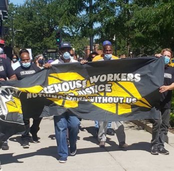 Warehouse workers march down a sidewalk wearing masks and holding a banner that says 
