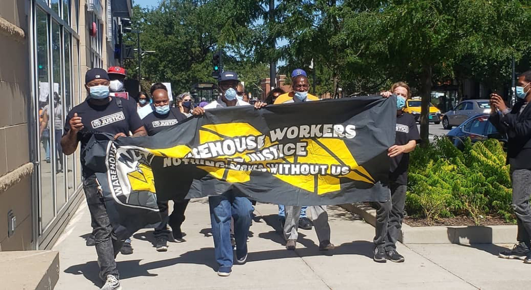 Warehouse workers march down a sidewalk wearing masks and holding a banner that says 