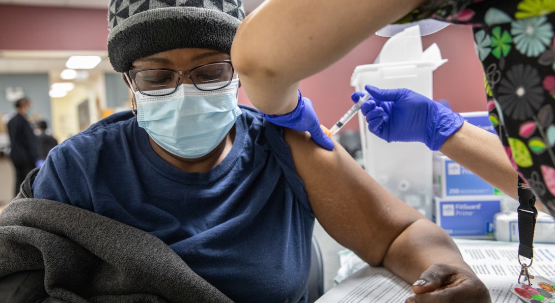 A nurse injects a COVID-19 vaccine into the arm of a female patient wearing a surgical mask.