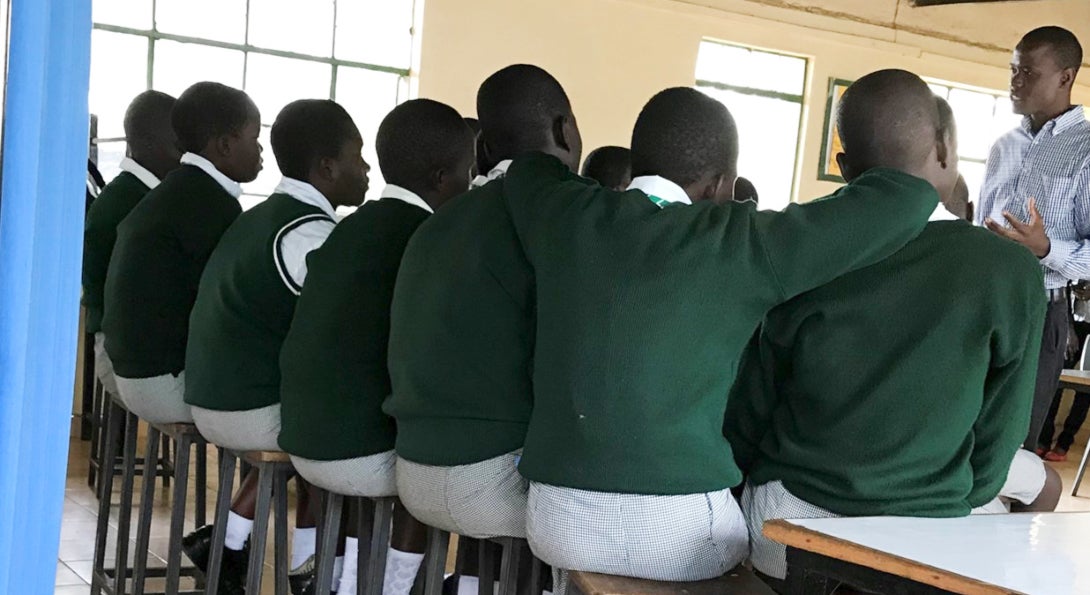 Students sit on stools listening to a talk from their teacher.