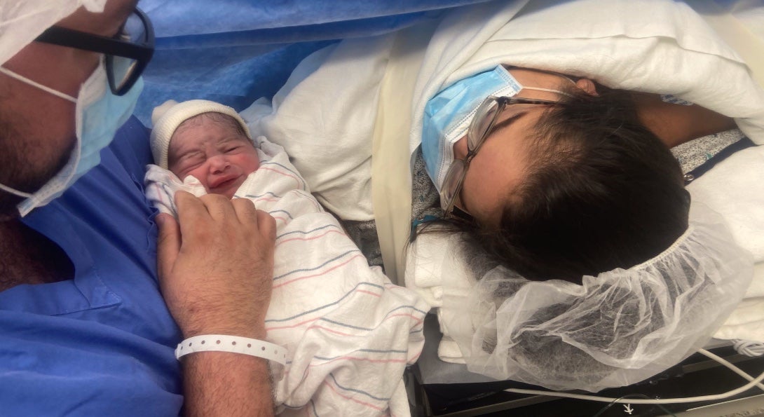 A father holds a newborn baby as the recuperating mother lies asleep in a bed nearby.