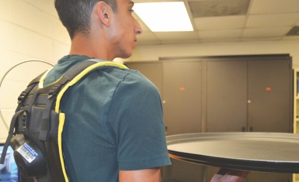A UIC student demonstrates a wearable AI sensor, a black packpack with sensors embedded inside. The student is holding a restaurant serving tray; the sensor monitors the student's posture as they are carrying a heavy tray.