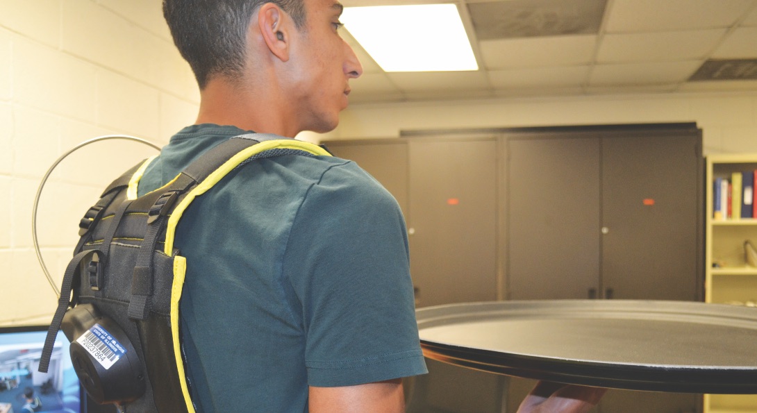 A UIC student demonstrates a wearable AI sensor, a black packpack with sensors embedded inside. The student is holding a restaurant serving tray; the sensor monitors the student's posture as they are carrying a heavy tray.