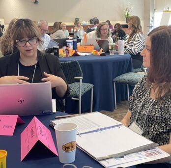 Two Leadership Institute Fellows discuss at a round table with paperwork and a computer with more fellows in the background at other tables 