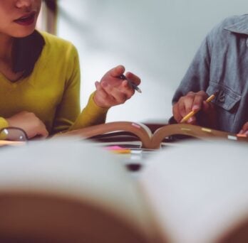 Two people sitting at a table with books open in front of them and glasses on the books. One person holds a pen in their hand and gestures towards the book.
                  