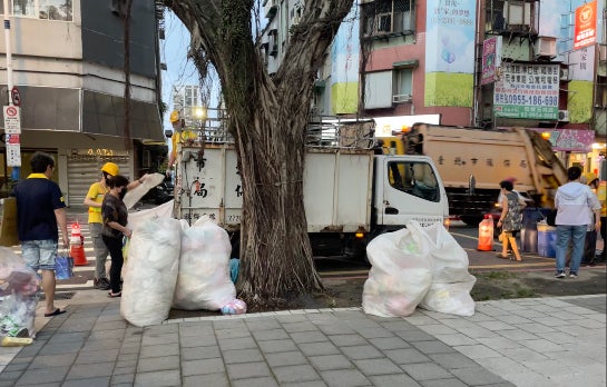 A white trash truck going down the street next to four white trash bags with people around.