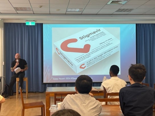 A person on a stool speaking to people with a screen showing a Powerpoint slide.